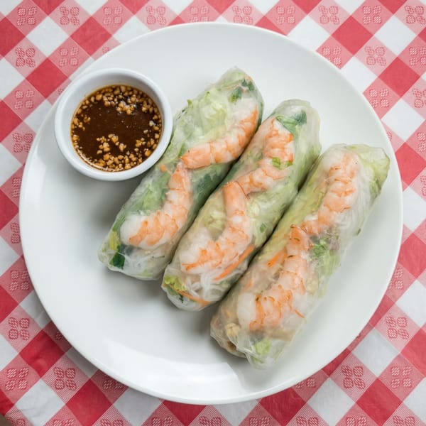 Three Thai Star shrimp spring rolls with peanut dipping sauce on a white plate and red checkered tablecloth.