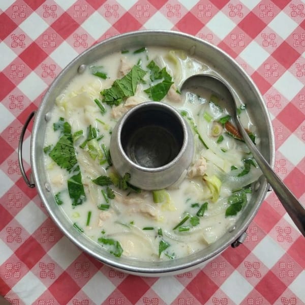 Creamy Thai soup with chicken, vegetables, and herbs in a metal hot pot on a red and white checkered tablecloth.