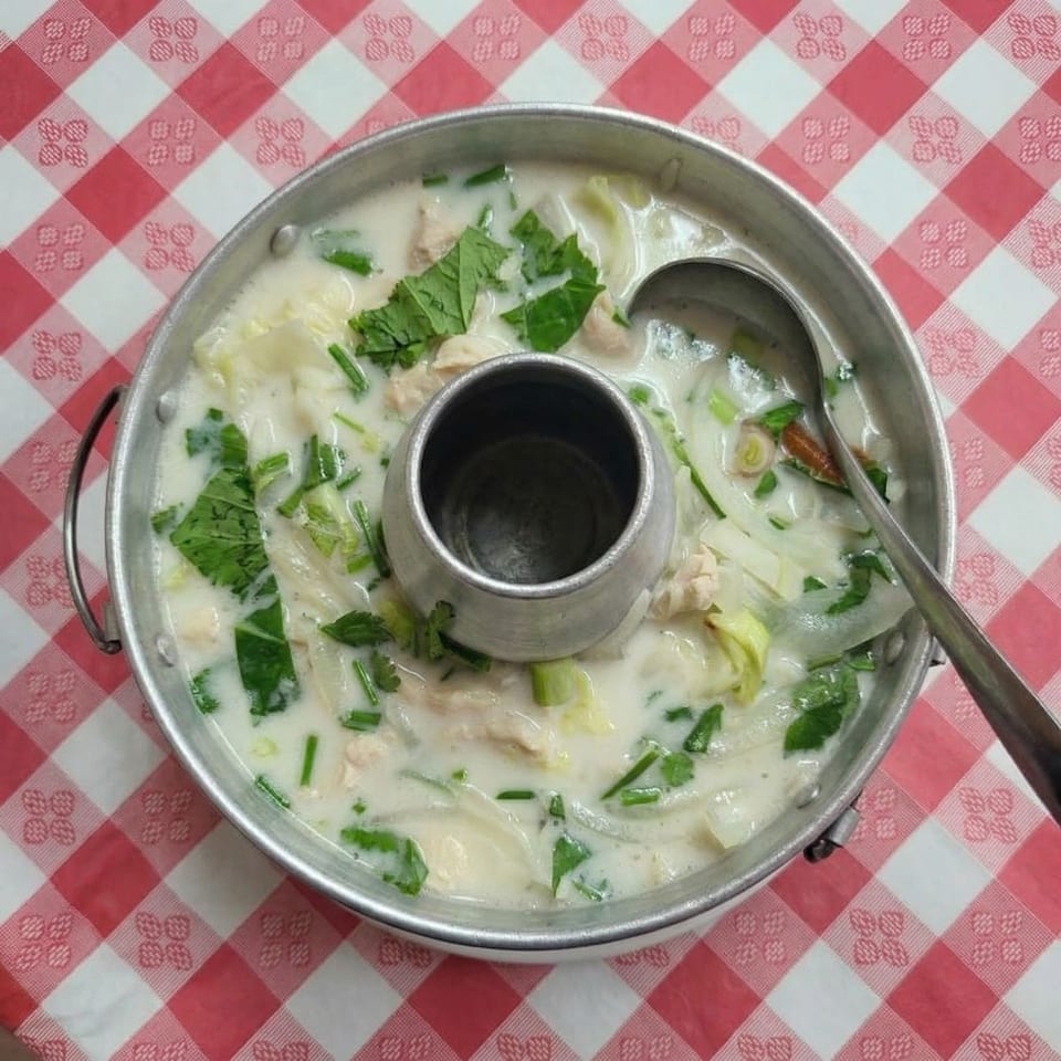 Creamy Thai soup with chicken, vegetables, and herbs in a metal hot pot on a red and white checkered tablecloth.